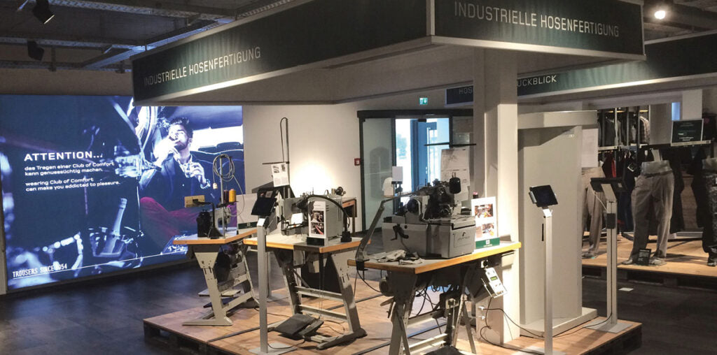 Ein Ausstellungsraum mit industriellen Maschinen und Geräten auf Tischen, einer beleuchteten digitalen Anzeige an der Wand, in der mehrere Personen an einem Messestand arbeiten oder sie durchsehen.