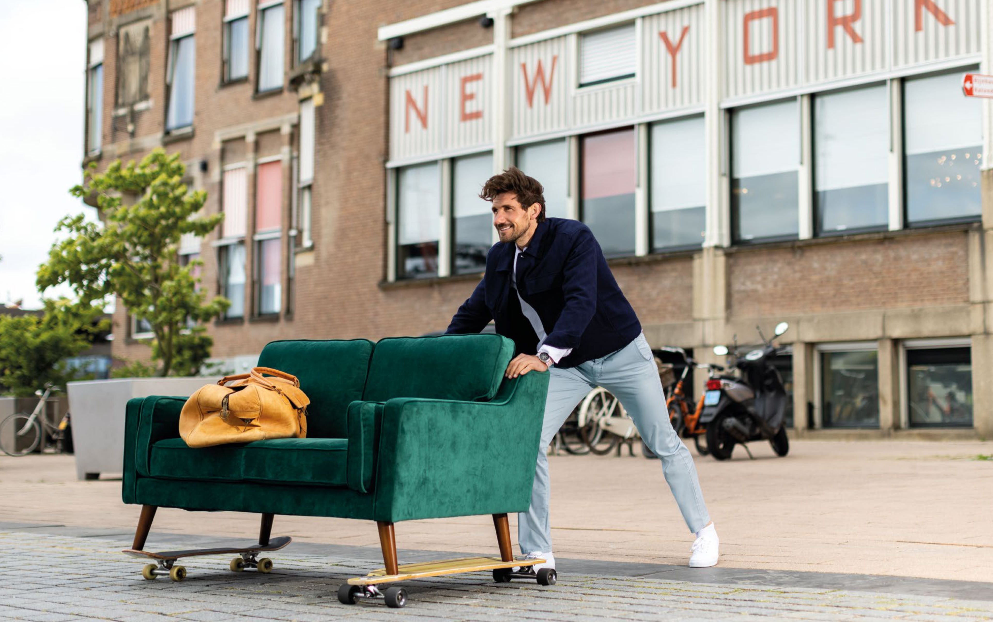 Ein Mann in einem blauen Blazer und weißen Hosen schiebt eine große grüne Couch mit Rollen vor einem Gebäude mit einer "New York"-Schriftzug. Auf der Couch liegt eine gelbe Tasche. Im Hintergrund sind Bäume, Fahrräder und ein Stadtgebiet sichtbar.