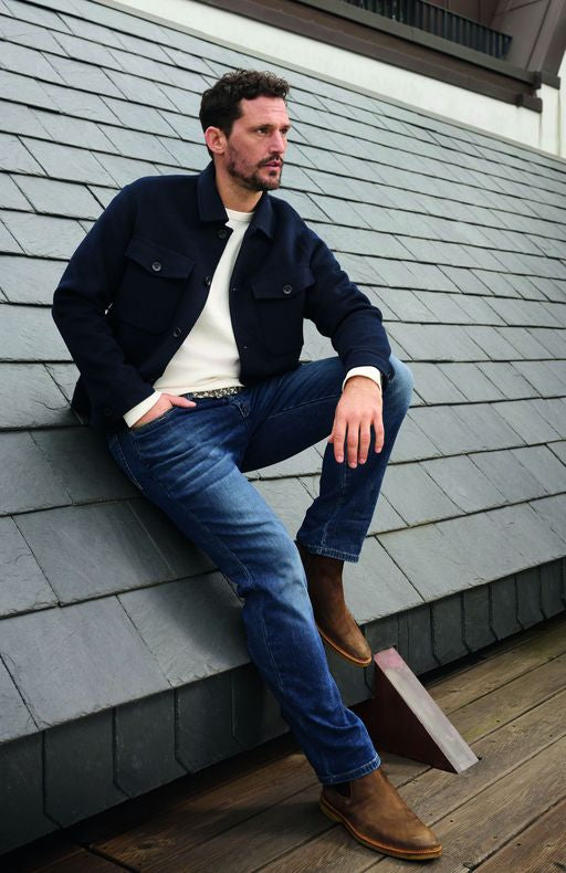 Mann sitzt entspannt auf einem Dach aus grauen Schindeln, trägt dunkelblaue Jeans, eine weiße Bluse, eine dunkle Jacke und braune Stiefel, schaut nach links und wirkt nachdenklich.