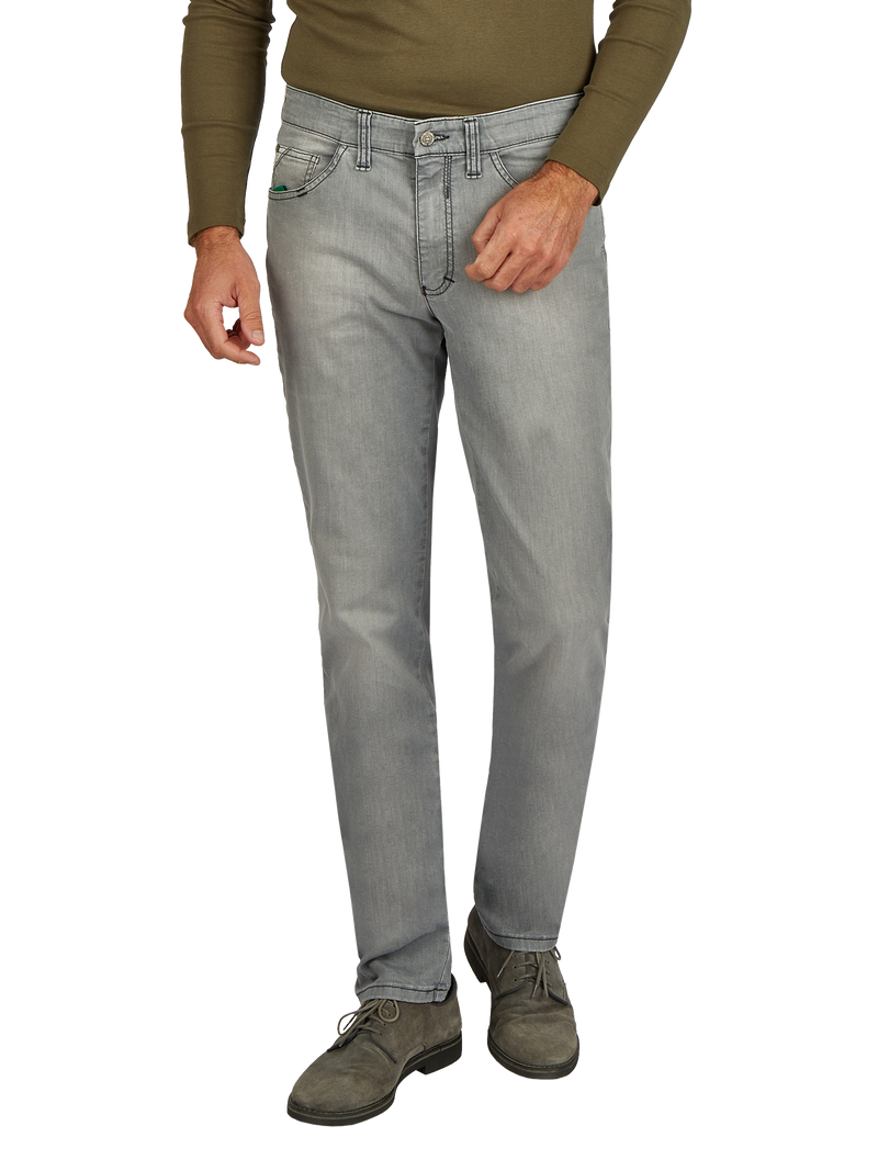 Der Bild zeigt einen Mann in einer grünen langärmligen Oberbekleidung und hellgrauen Jeans. Er steht mit einer Hand in der Hosentasche und trägt braune Schuhe. Das Bild fokussiert auf den unteren Bereich des Körpers.