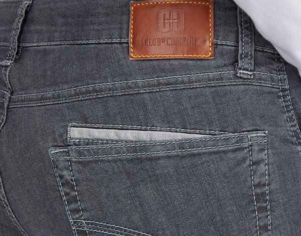 Nahaufnahme der Rückseite eines schwarzen Jeanshosen mit einer braunen Lederlasche am oberen Rand, auf der das Logo "CLUB & COMPLICE" und ein Kreuzsymbol geprägt sind. Die Jeans zeigt eine hintere Tasche mit Doppelnaht.