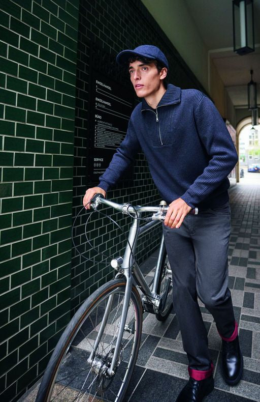 Ein junger Mann mit schwarzem Haar, weißen Hautfarbe und braunen Augen steht mit einem silbernen Fahrrad vor einer schwarzen Wand mit grünen Fliesen. Er trägt eine blaue Mütze, einen blauen Zopf, eine blaue Fleece-Jacke mit Reißverschluss, dunkle Hosen, schwarze Schuhe mit roten Akzenten und schaut nach rechts.