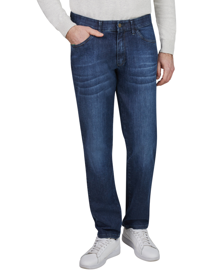 Mann trägt blaue Jeans, die vorne die linke Hand in der Tasche, trägt weißes T-Shirt und weiße Sneakers, während er links steht.