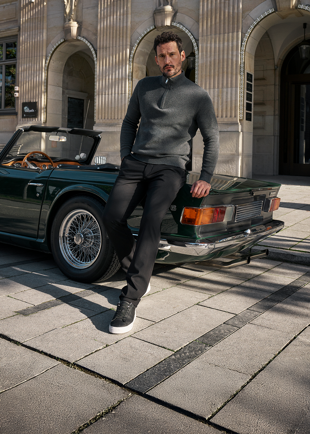Junger Mann mit dunklem, kurz curly Haar, dunklem Bart und ernstem Gesichtsausdruck lehnt entspannt an einem schwarzen Oldtimer-Cabriolet auf einer Stadtstraße vor einem historischen Gebäude mit großen Bögen und leuchtender Beleuchtung.