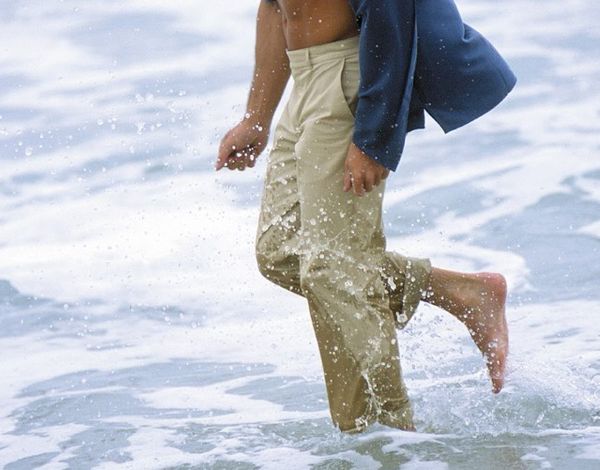Ein Mensch, nur teilweise im Bild sichtbar, läuft barfuß im Wasser am Strand. Die Person trägt beige Hosen und ein blaues Oberteil. Die Wellen umspülen die Füße und erzeugen kleine Spritzer.