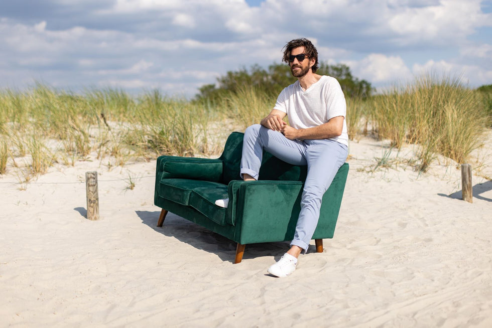 Ein Mann mit Sonnenbrille sitzt entspannt auf einem grünen Samtsofa inmitten von Sanddünen und Gras, passend zum dunkelgrünen Farbton des Club of Comfort Sofas.