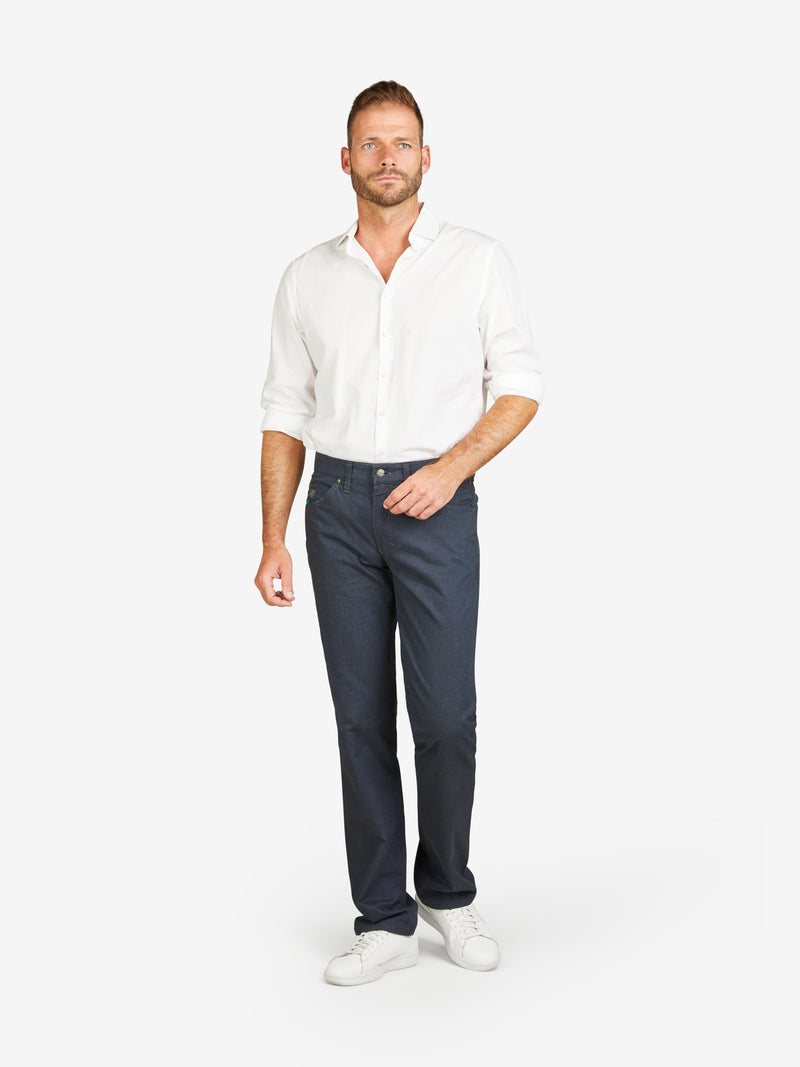 Mann trägt dunkelblaue HENRY 6910 5-Pocket-Hose aus Baumwoll-Mikrostruktur mit Wool-Look, elastischem Doppelstoffbund, Mobile Pocket in rechter Vordertasche, Kontrast-Münztasche, normaler Leibhöhe, schmalem Bein, weißes Hemd und Sneakers, neutraler Hintergrund.