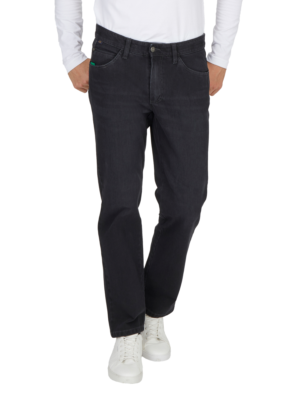 Ein Mann trägt dunkle Jeans, die locker sitzen, und eine weiße Langarmbluse. Seine Hände sind in den Taschen der Jeans. Er trägt weiße Sneakers und steht vor einem weißen Hintergrund.